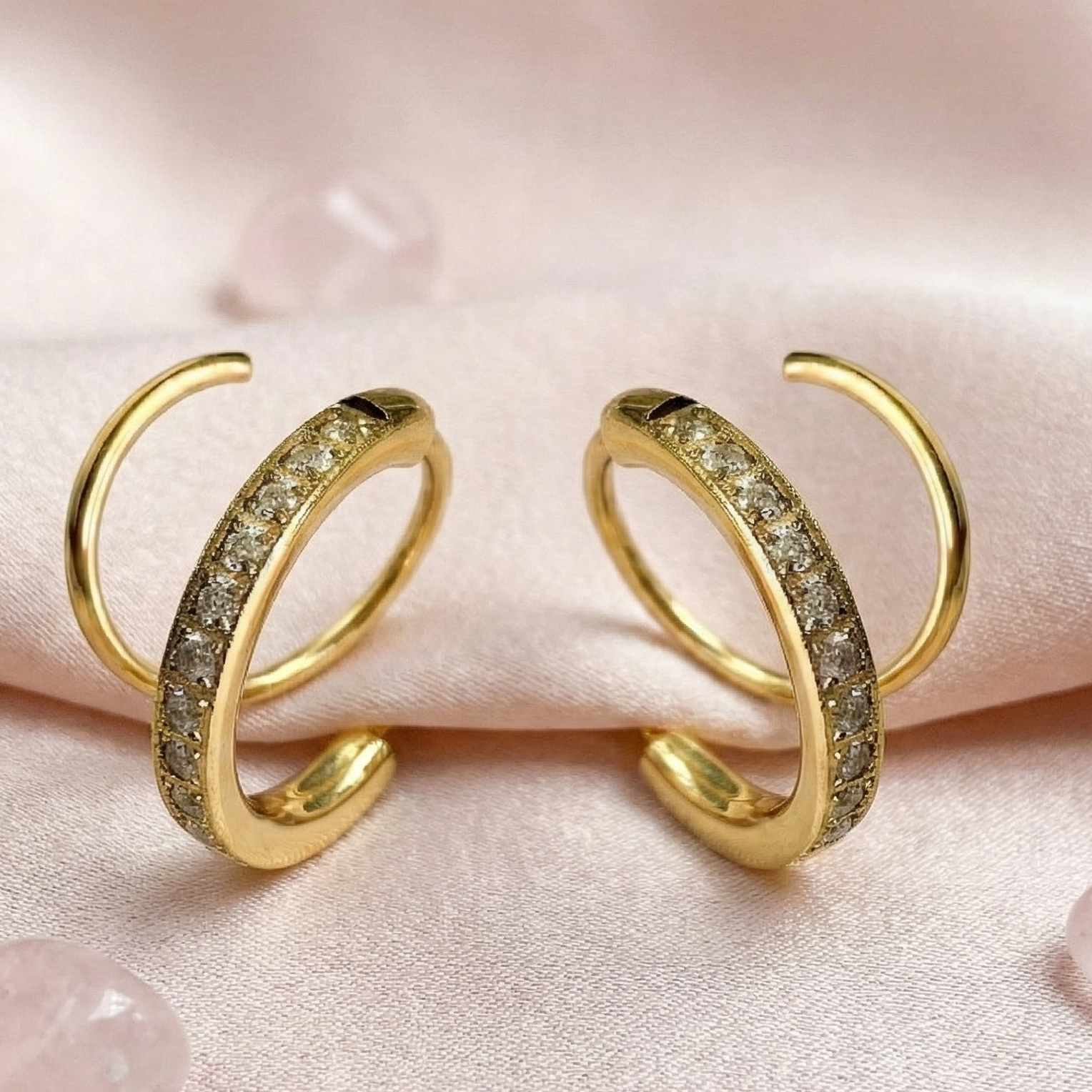 Gold hoop earrings with embedded stones on a soft pink fabric background